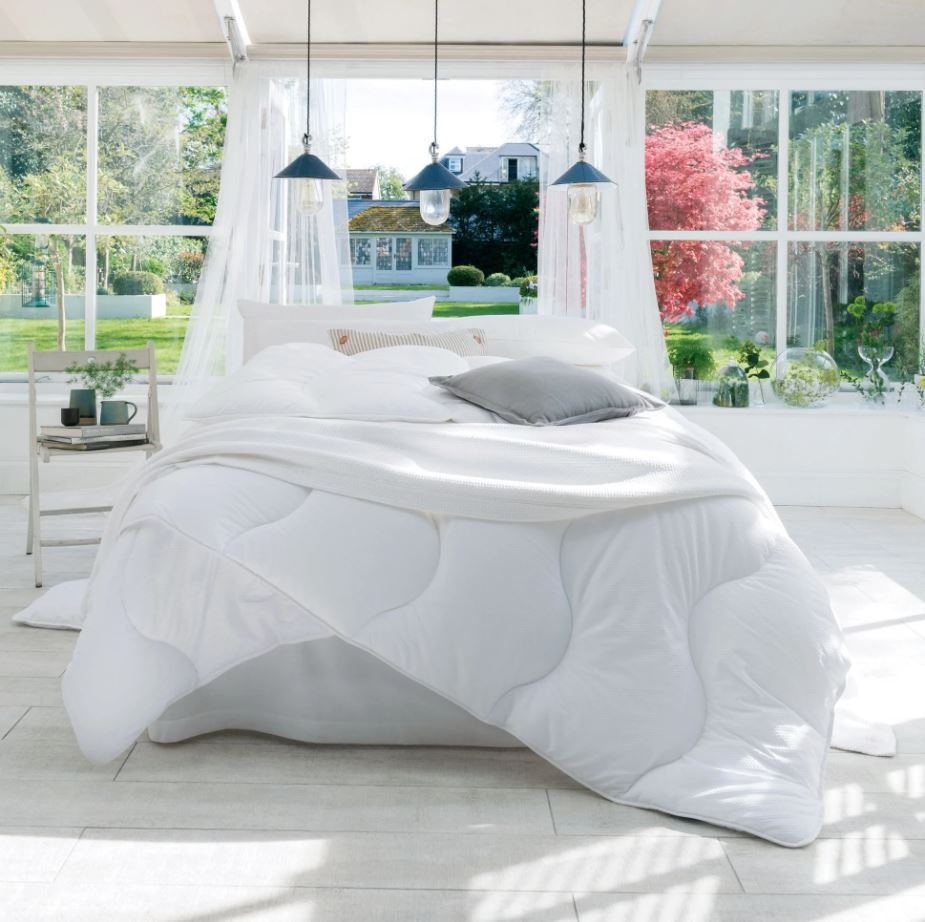 Bed with a white duvet and pillows in the center of a bright room with big windows onto a garden